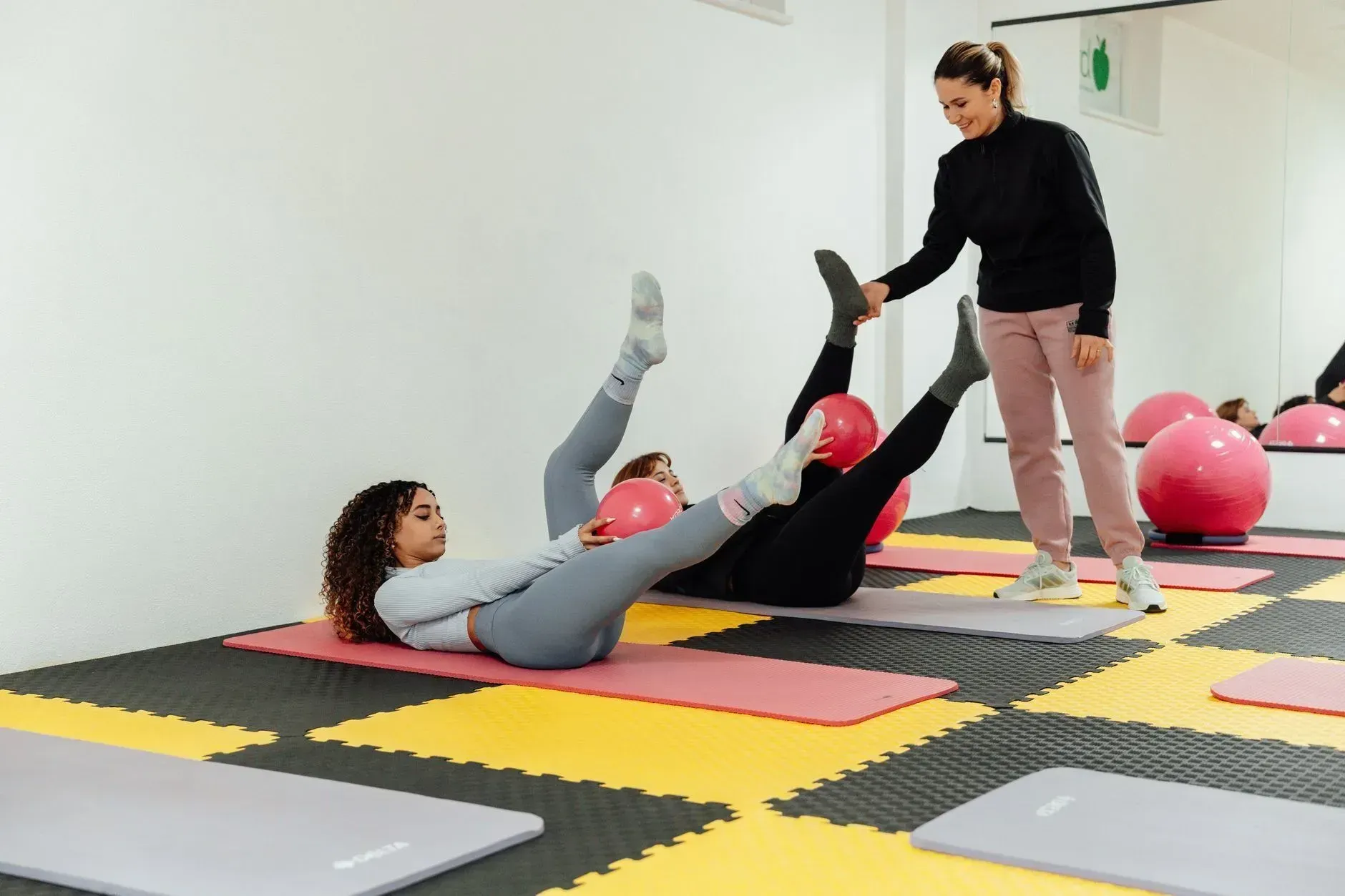 Three people exercising on mats in a gym, doing leg lifts with pink exercise balls nearby