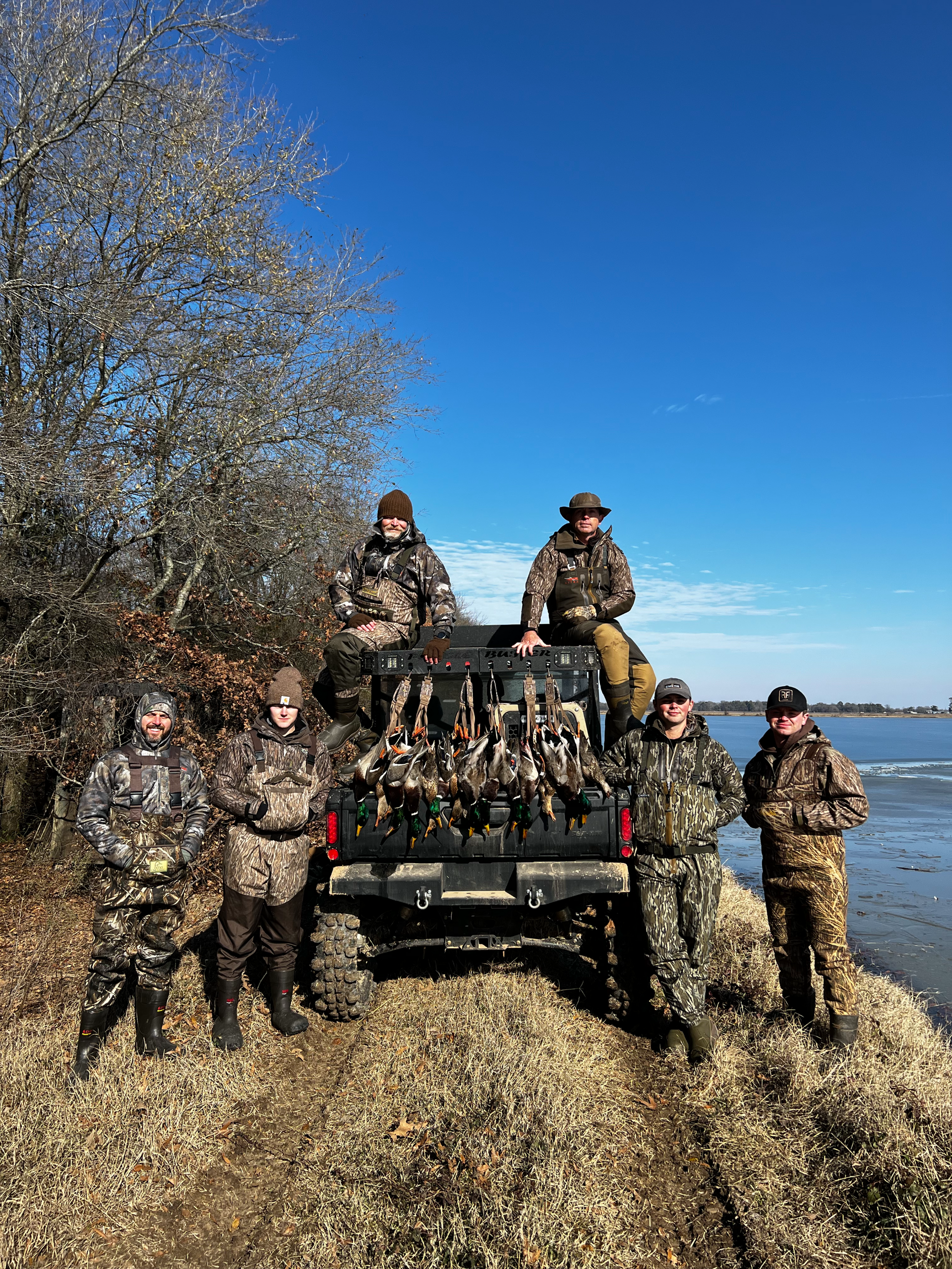 People in camouflage posing on an ATV in a leaf-covered field under a clear blue sky.
