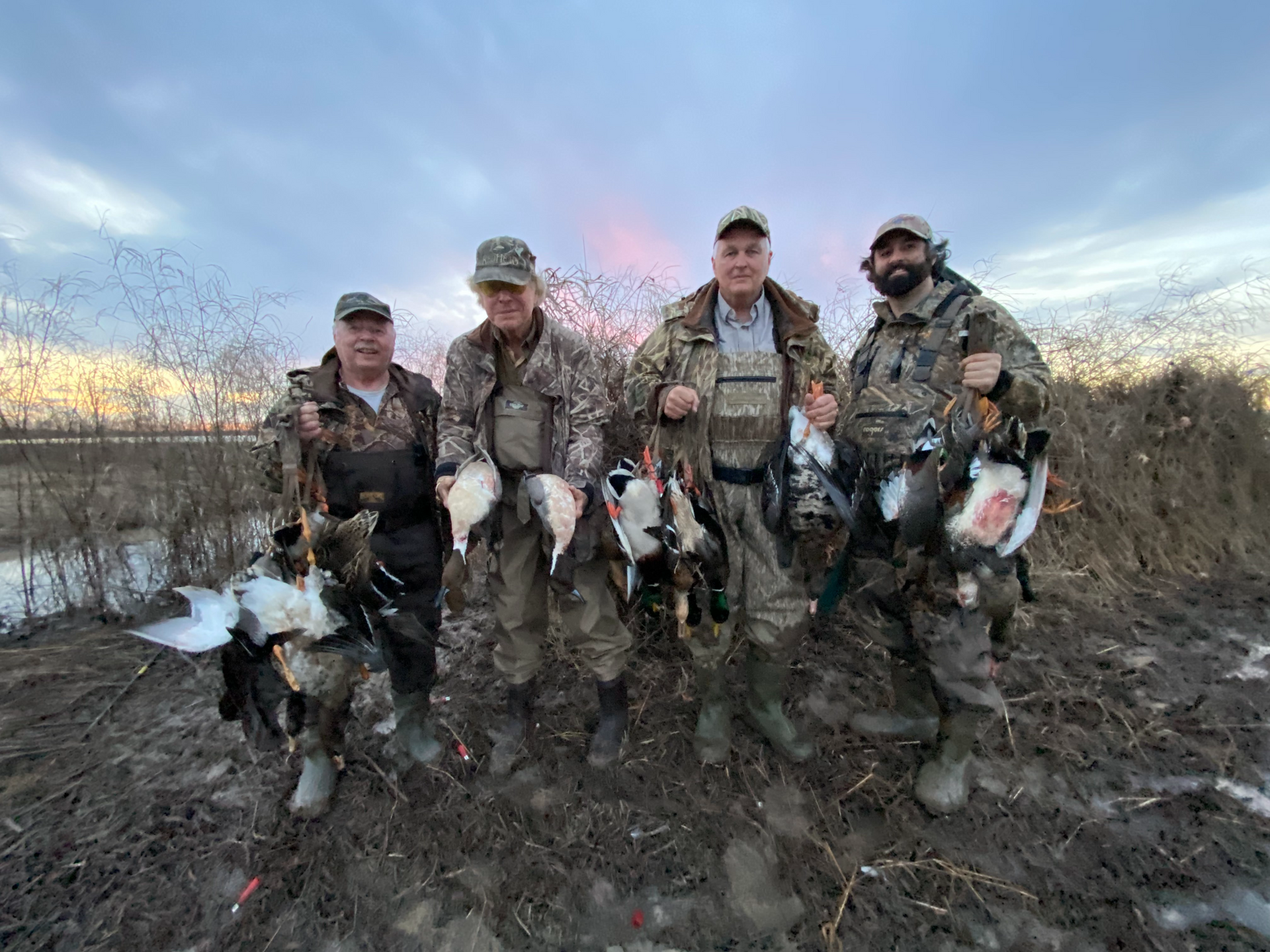 Four hunters in camouflage pose with several dead geese in a muddy wetland at dusk