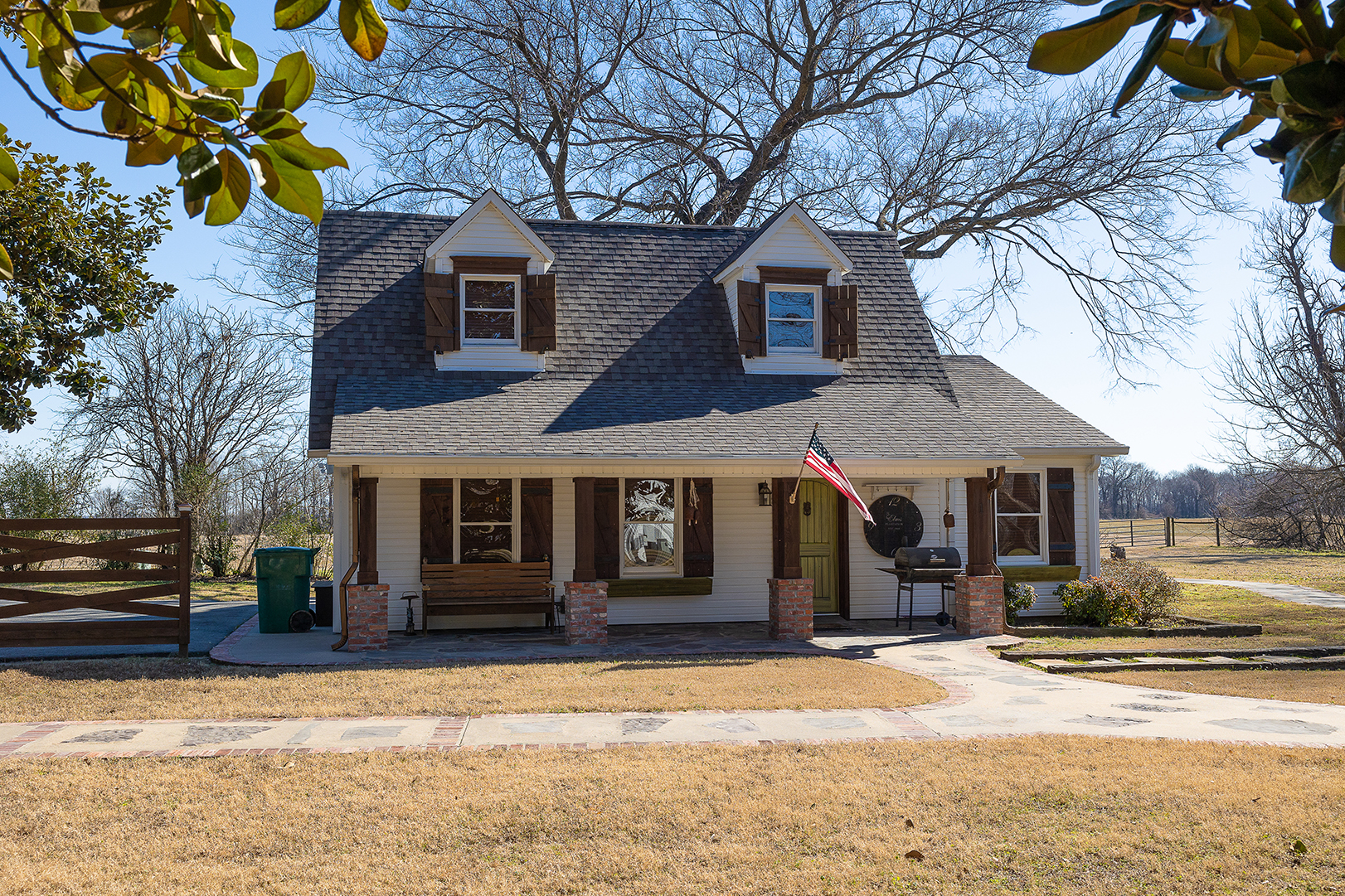 Two-story farmhouse with a porch, brick chimney, and American flag on a sunny rural lot