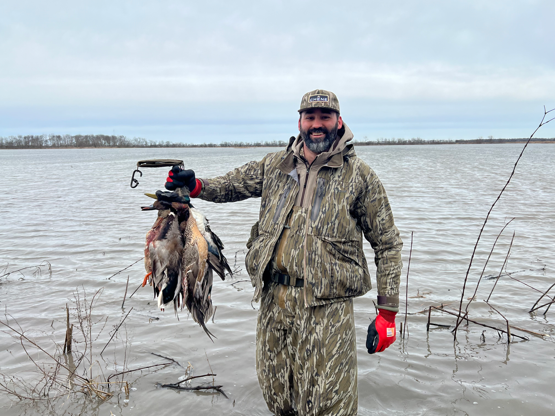 Camouflaged hunter holding several birds by a frozen lakeshore under a cloudy sky