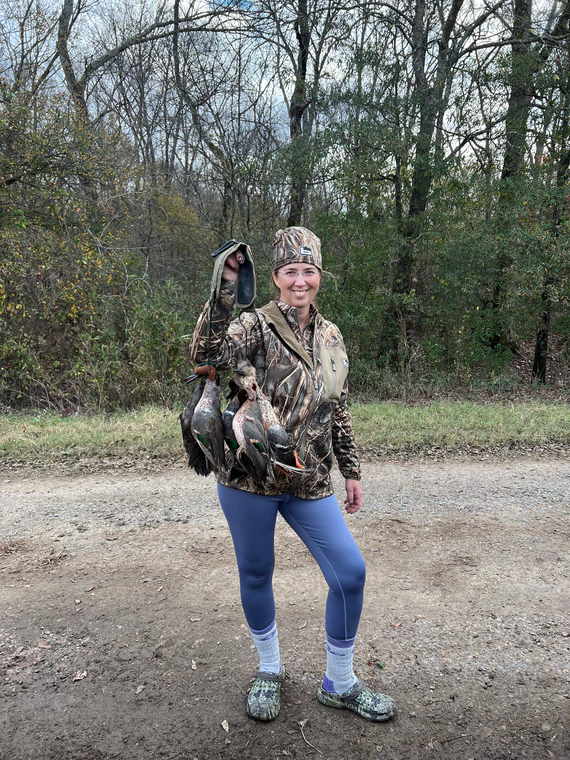 Person in camouflage holding a fish on a gravel road beside trees.