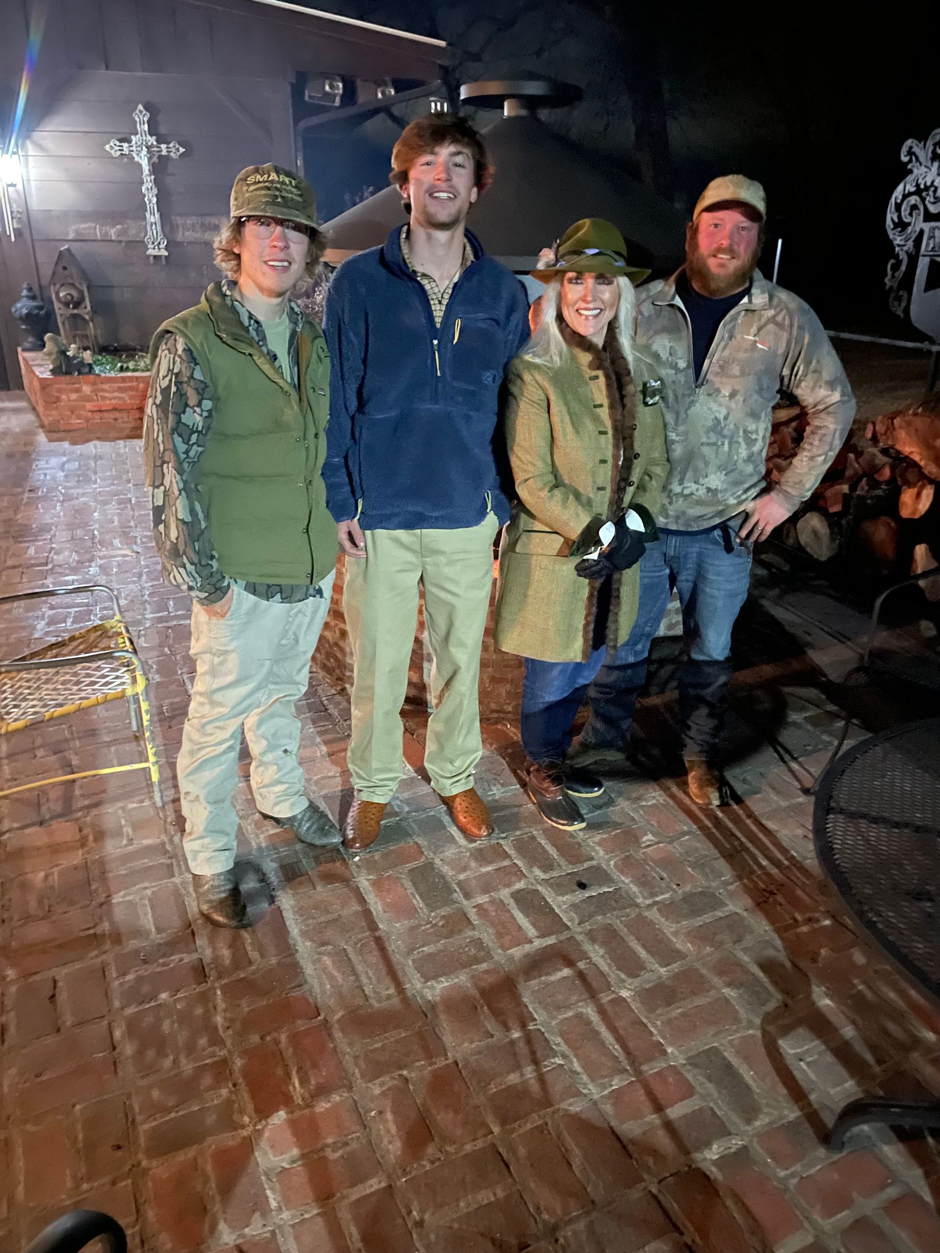 Four people in outdoor gear stand on a brick patio at night, smiling near a lit building.