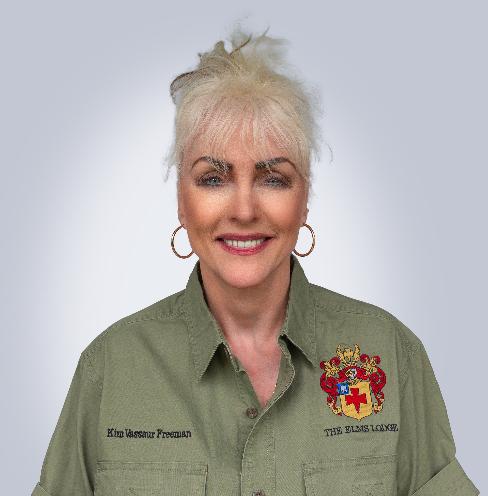Woman smiling in olive green shirt with embroidered crest, against a plain light background.