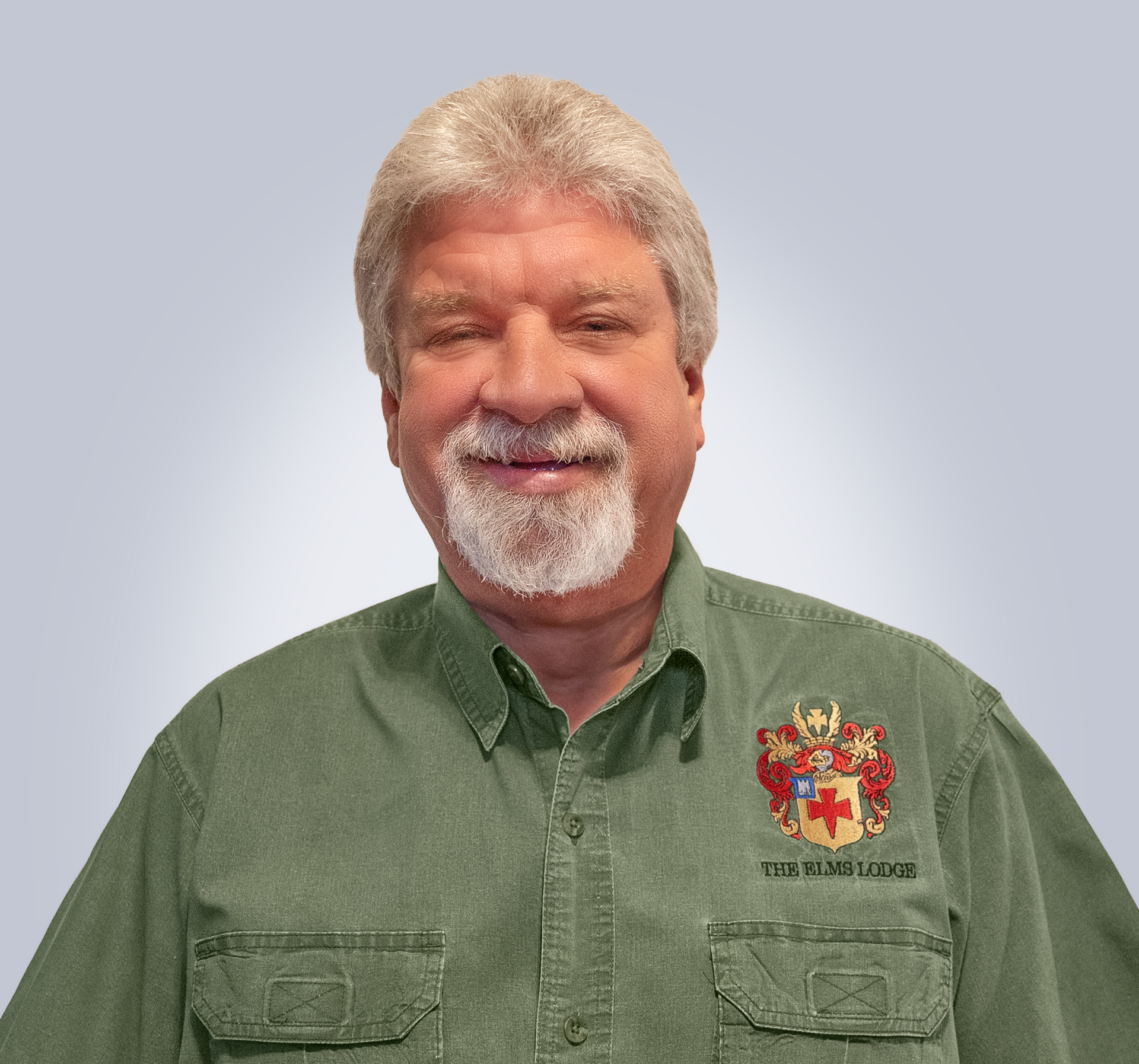 Smiling man in green button-up shirt with embroidered crest, standing against a plain gray background