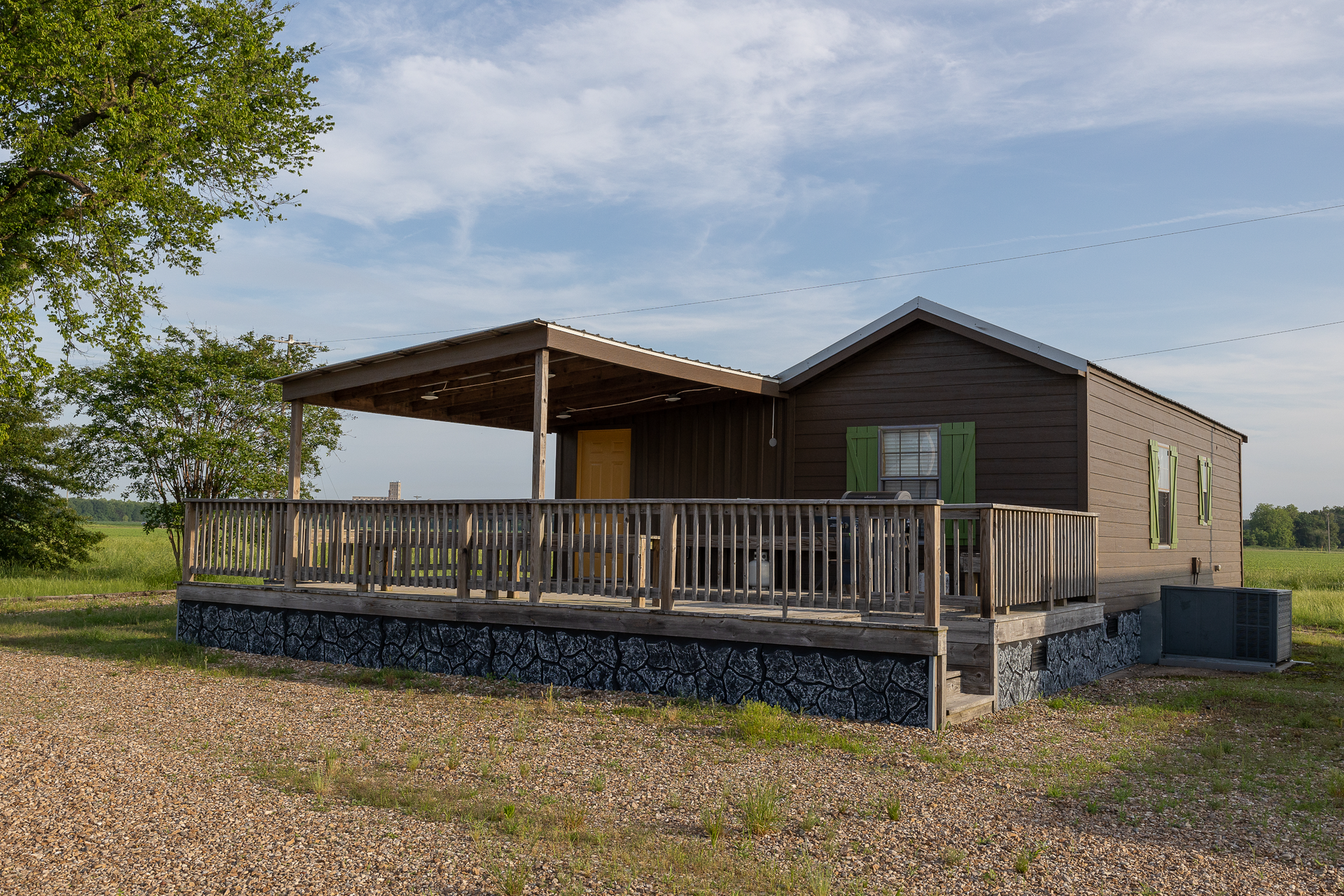 Small tan cabin with a covered porch and wooden railing in a grassy rural field