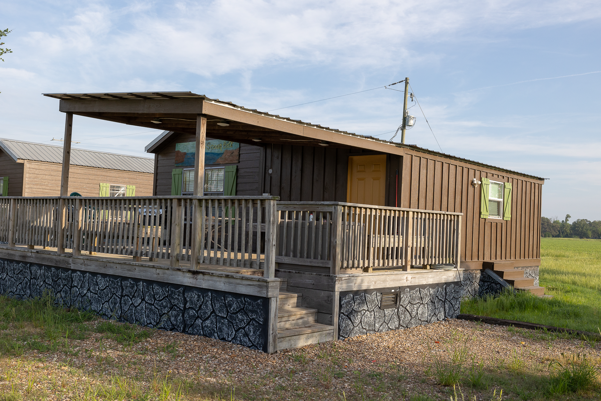 Small wooden cabin with a covered porch and railings on a grassy lot under a blue sky.