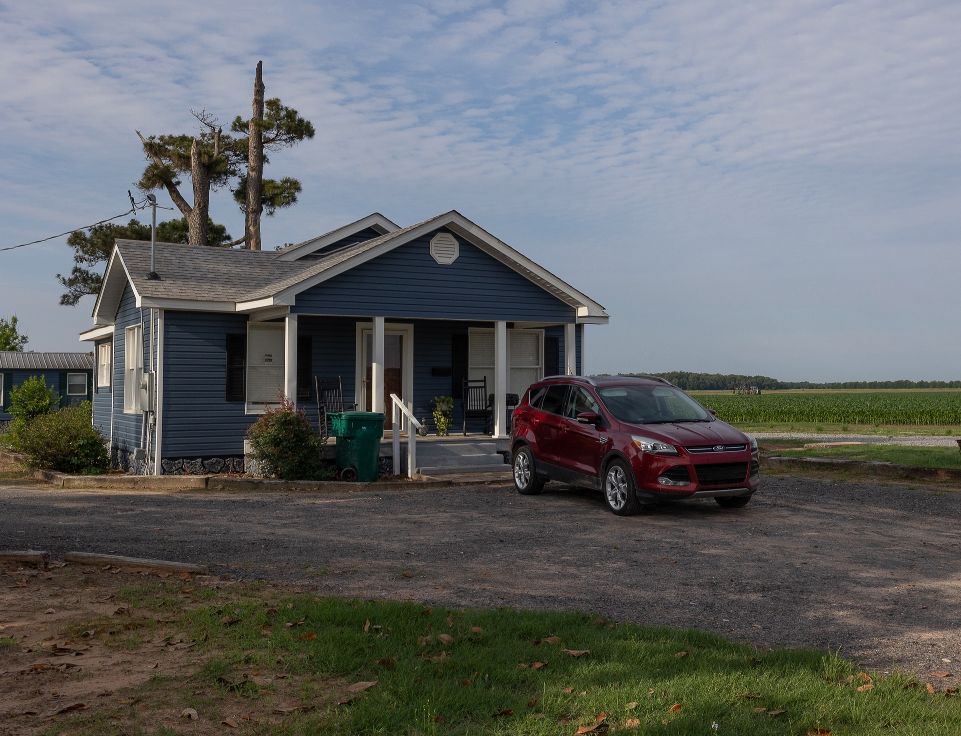 Small blue house by a rural road with a red SUV parked in front, open fields in the background.