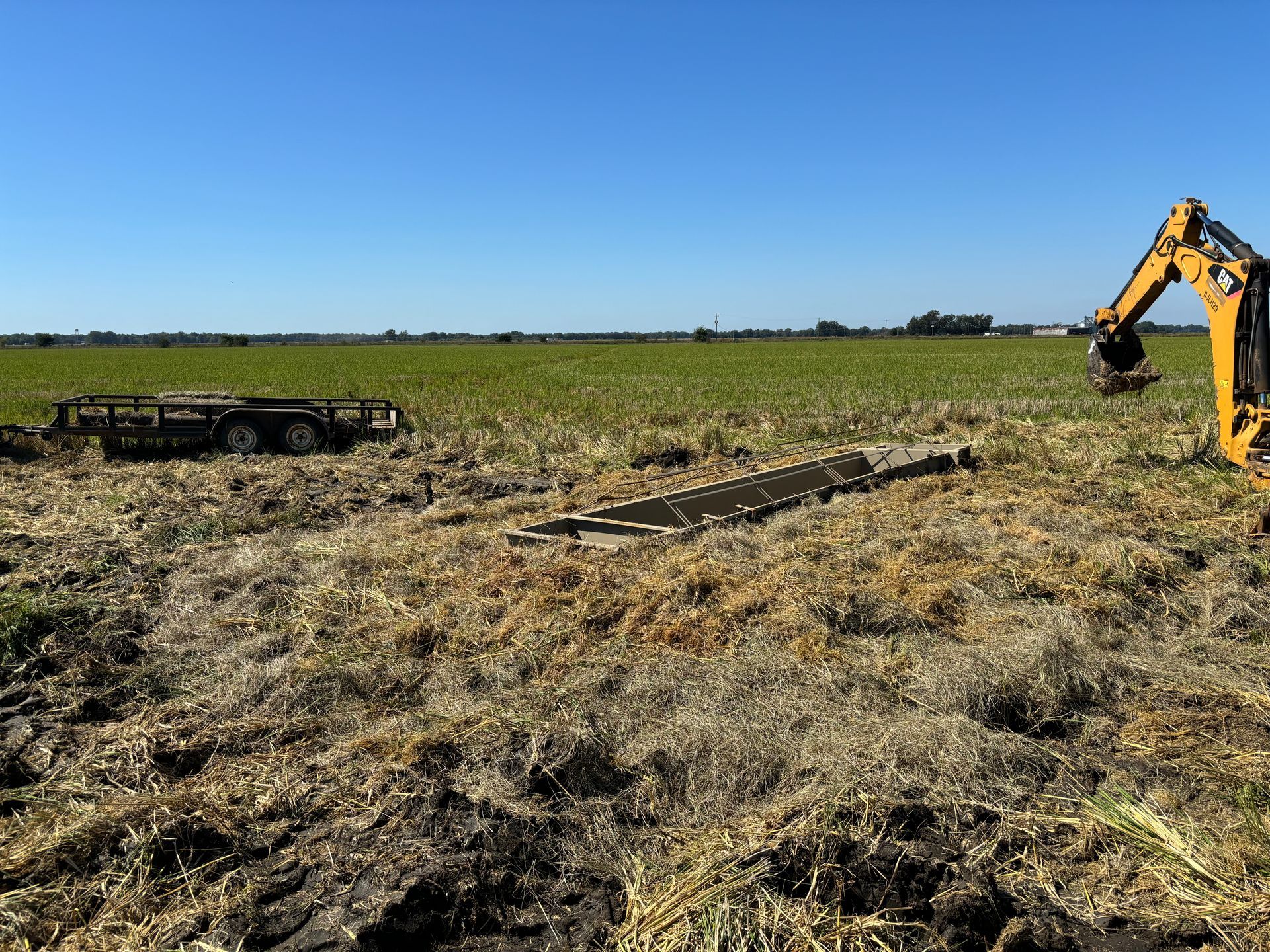 A yellow excavator works in a muddy field under a clear blue sky.