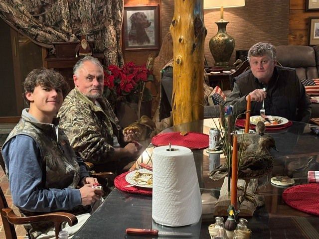 Three people seated at a dining table with plates, candles, and a paper towel roll in a cozy room