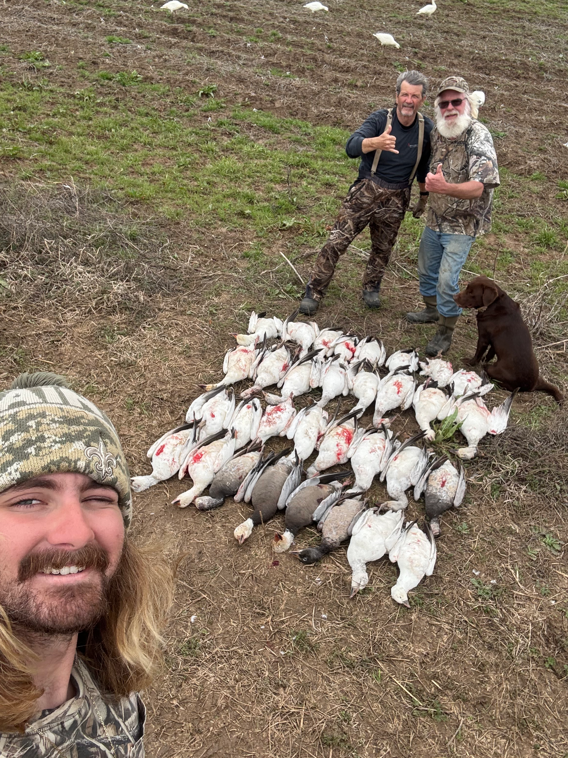 Three people posing outdoors beside a large pile of harvested birds, with a dog nearby.