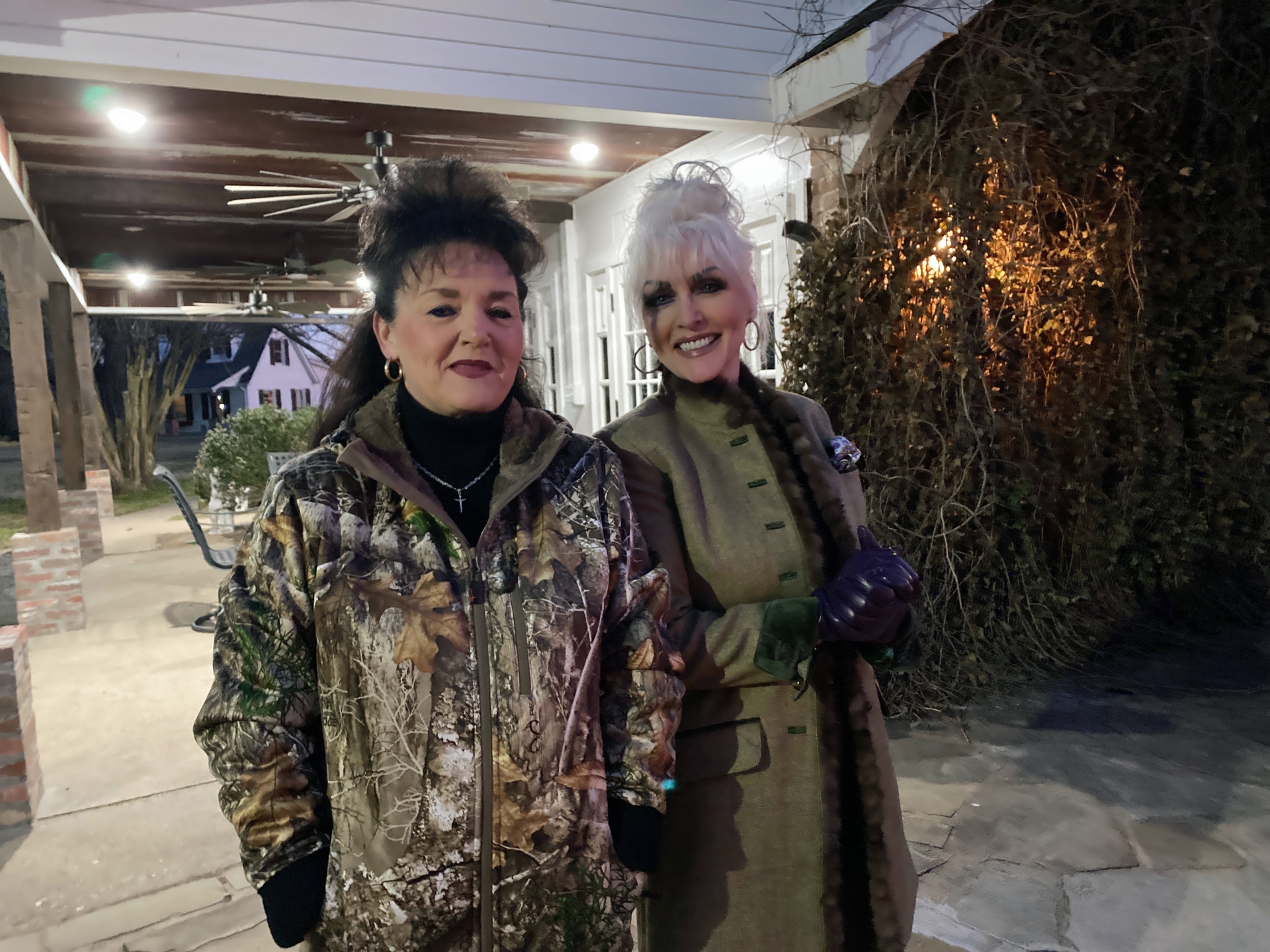 Two women smiling outdoors at night, standing under a covered walkway with string lights.
