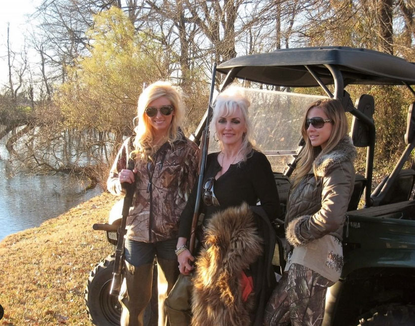 Three people posing beside an off-road vehicle by a wooded riverside in autumn sunlight