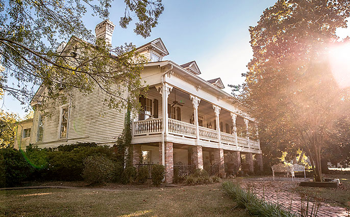Historic white house with wraparound porch, trees, and sunlight filtering through the yard