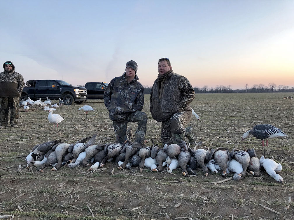Two hunters in camouflage stand behind a row of geese on a field at sunset.