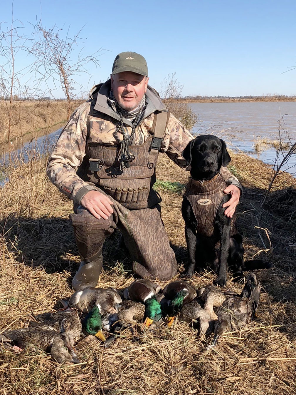 Hunter kneeling by a black dog beside several ducks in a marshy field