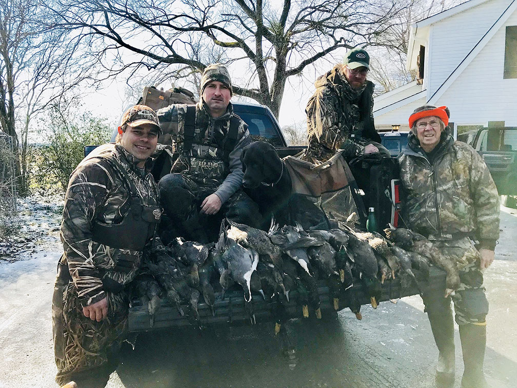 Four hunters posing with a deer in the back of a truck on a snowy street