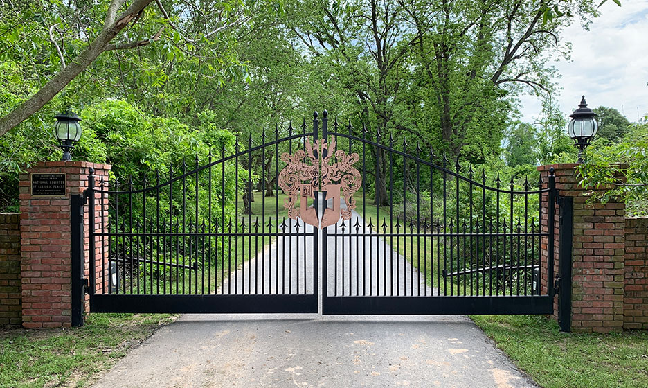 Black wrought-iron gate with brick pillars at a leafy driveway entrance