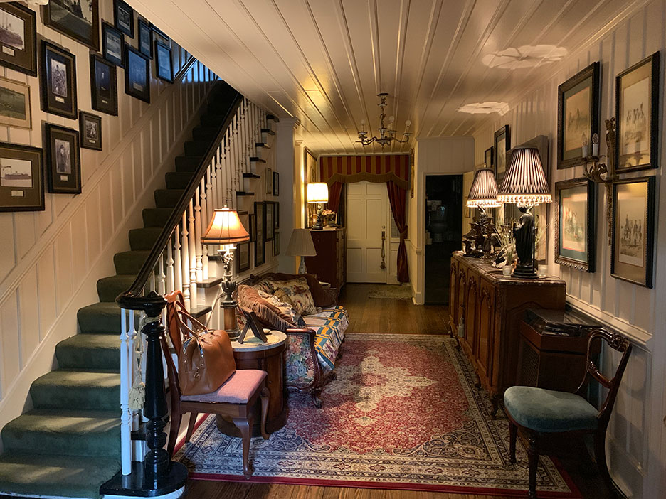 Cozy hallway with staircase, framed photos, vintage furniture, patterned rug, and warm lamp lighting