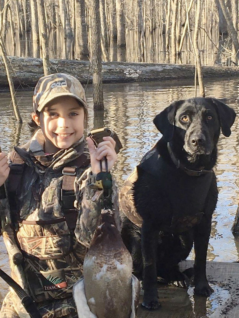 Person in camouflage holding a duck beside a black dog by a lakeside during a hunt