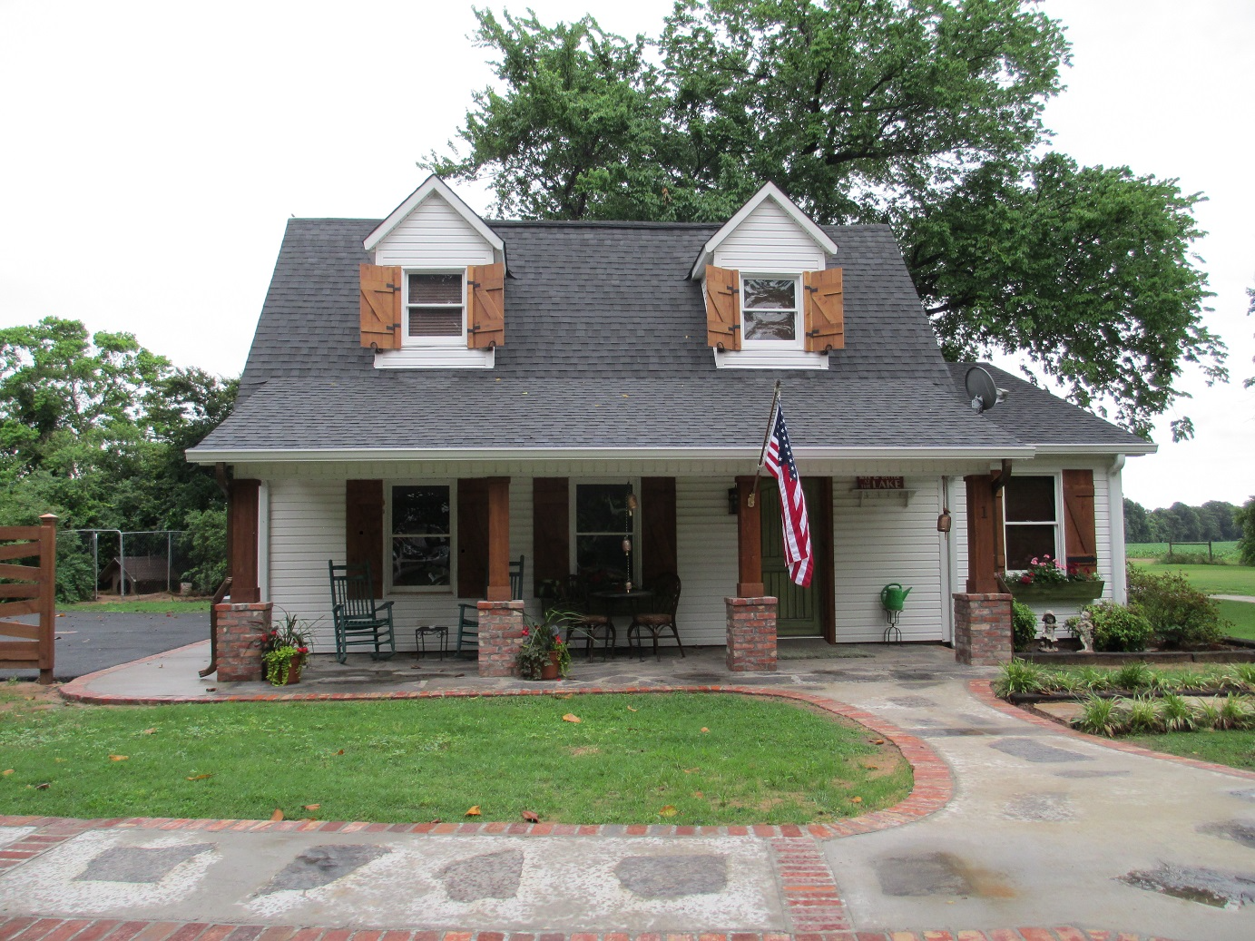 Small white house with dark roof, brick porch, American flag, and front yard landscaping.