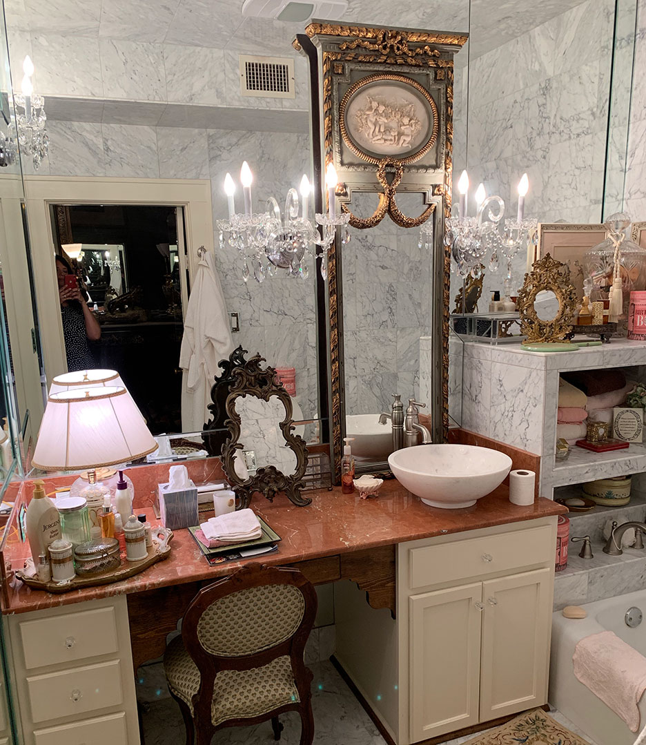 Vanity in a marble bathroom with ornate mirror, pink lamp, and white sink under chandelier sconces