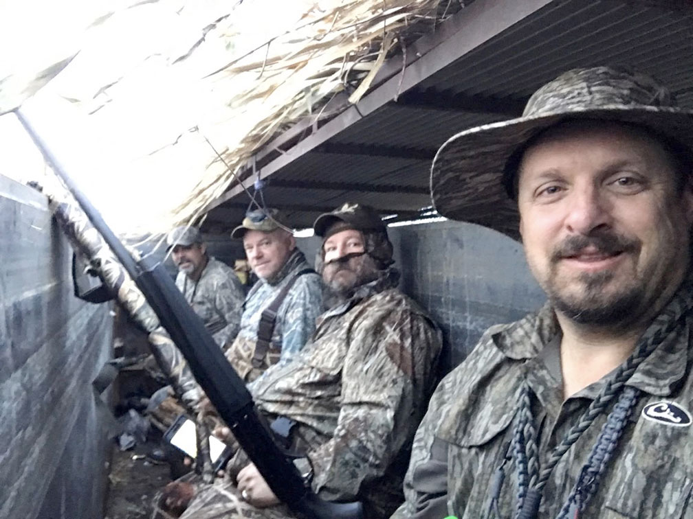 Four hunters in camouflage gear sit in a blind, holding rifles and looking toward the camera.