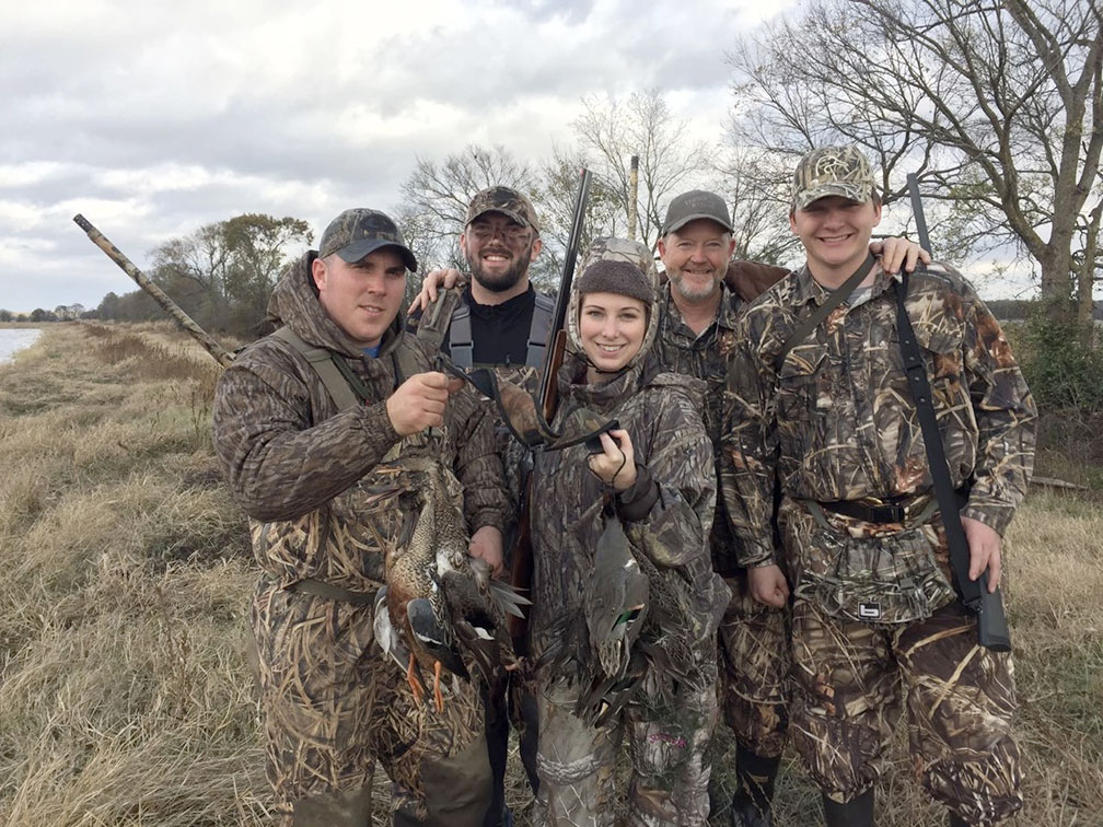 Five hunters in camouflage pose with game beside a marshy field.