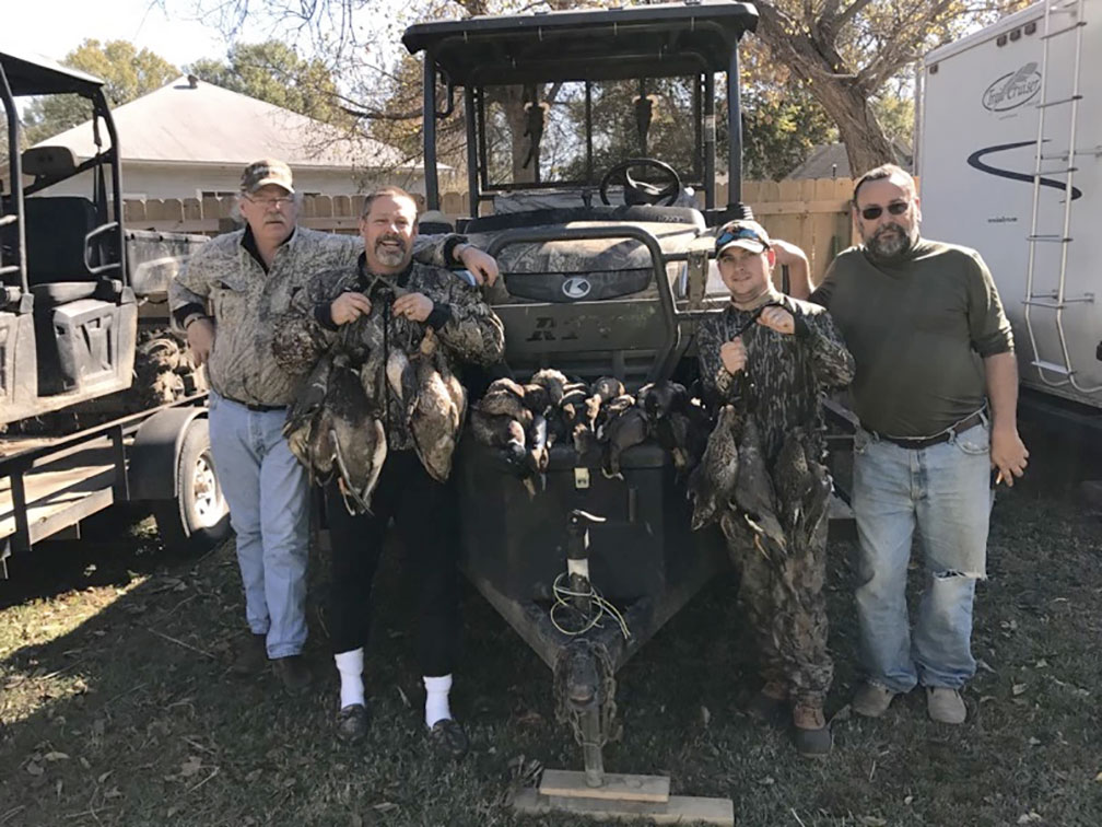Four people posing with hunting gear and a utility vehicle outdoors, holding game birds.