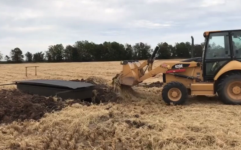Construction vehicle moving dirt near a black tarp in a harvested field under a cloudy sky