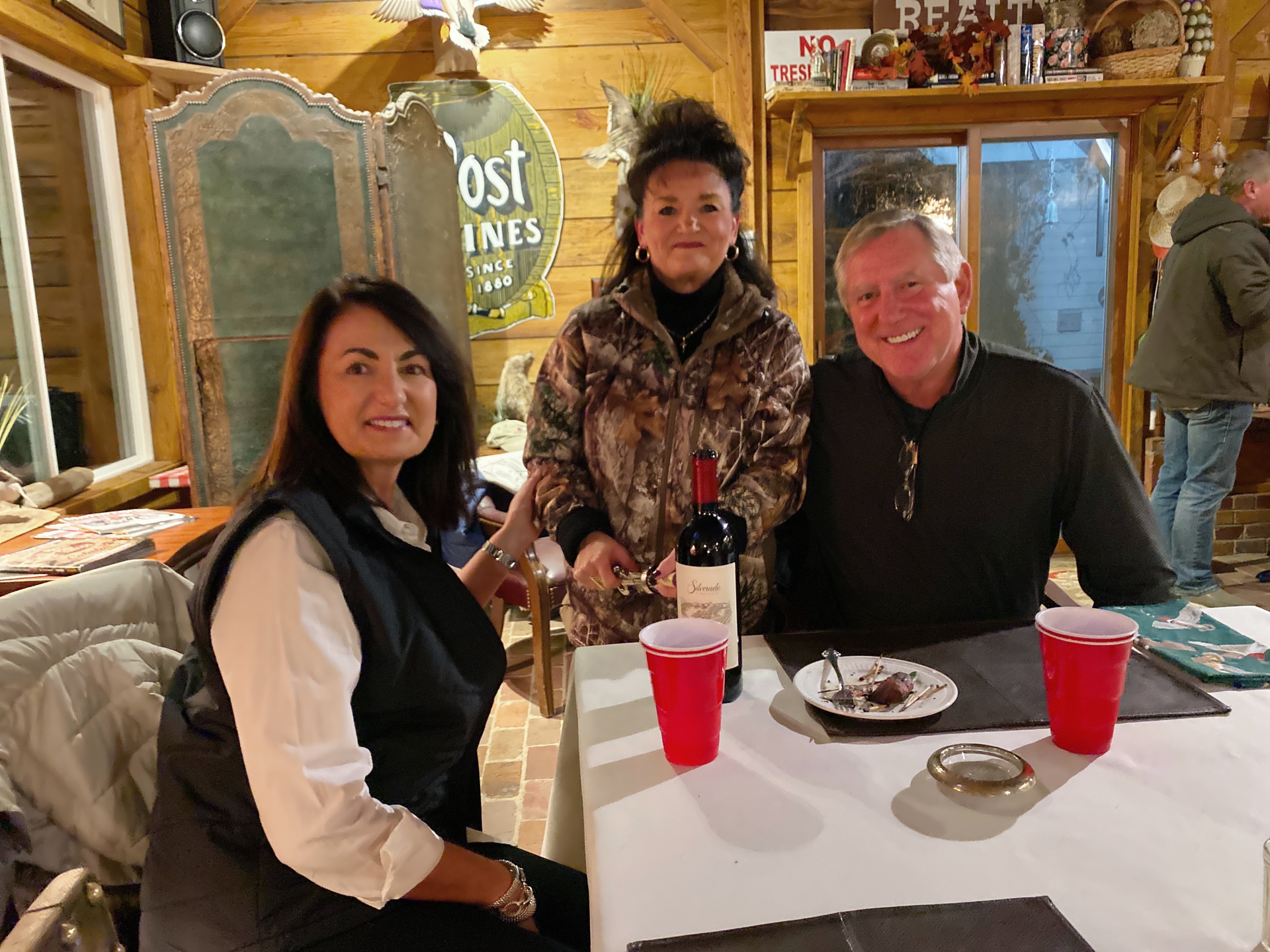 Three people smiling at a restaurant table with drinks and dessert in a cozy rustic interior.