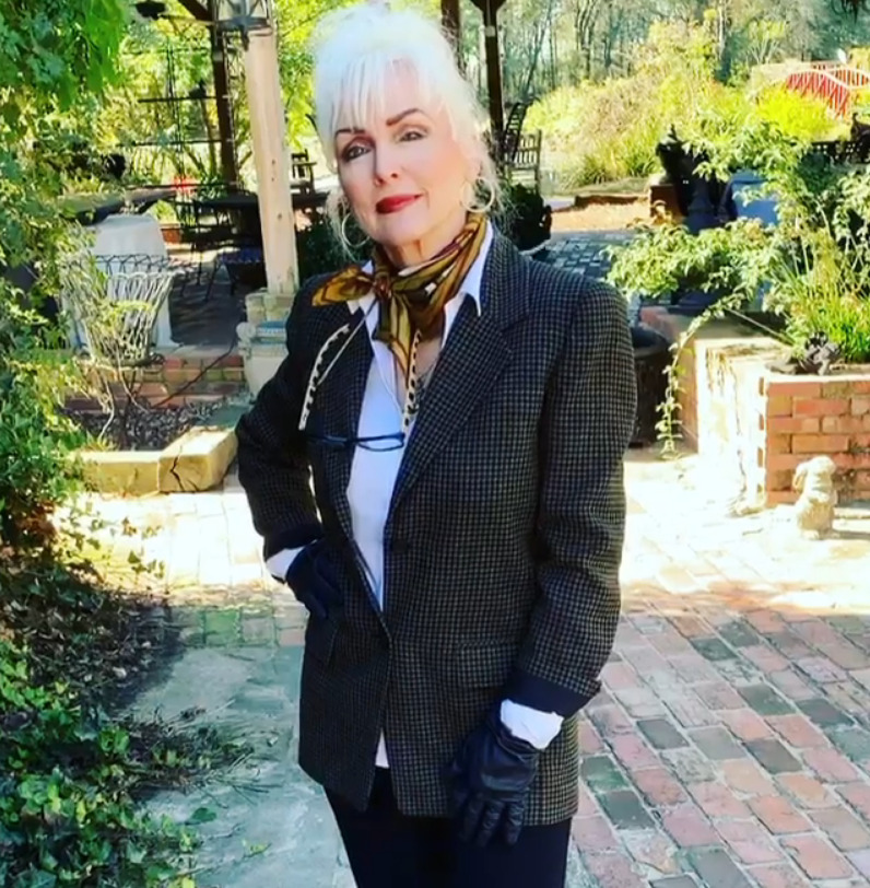 Woman standing on a garden path in a dark blazer, patterned scarf, and white blouse, with greenery and brick behind her