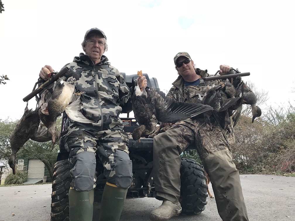 Two hunters in camouflage pose with a large deer on an ATV, outdoors on a road.