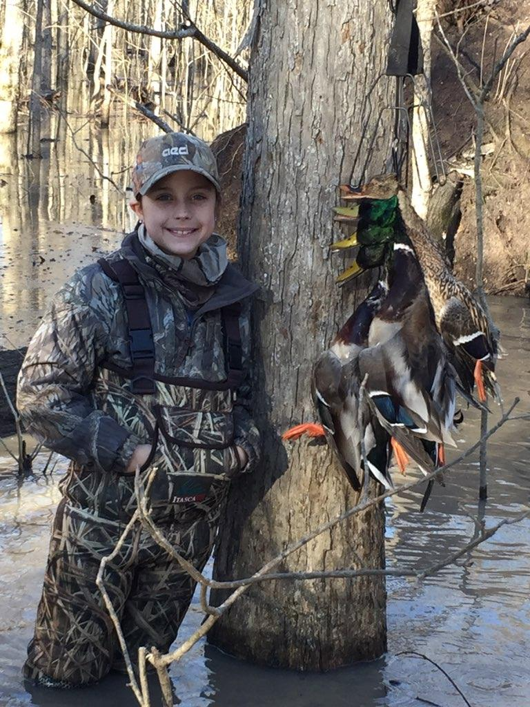 Person in camouflage holding ducks beside a tree in shallow water.