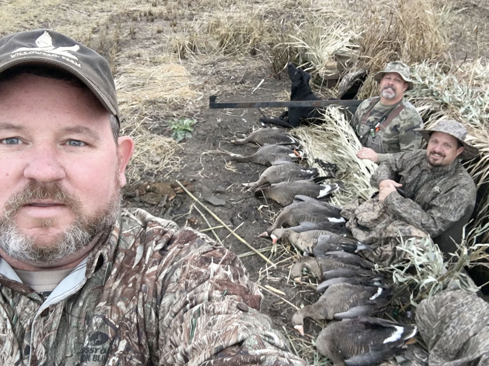 Three hunters pose with a row of ducks in a grassy field after a hunt