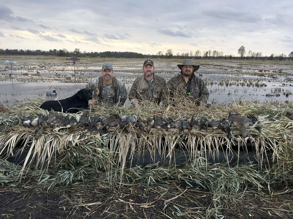 Three hunters in camouflage kneel behind a blind in a marsh with waterfowl decoys.