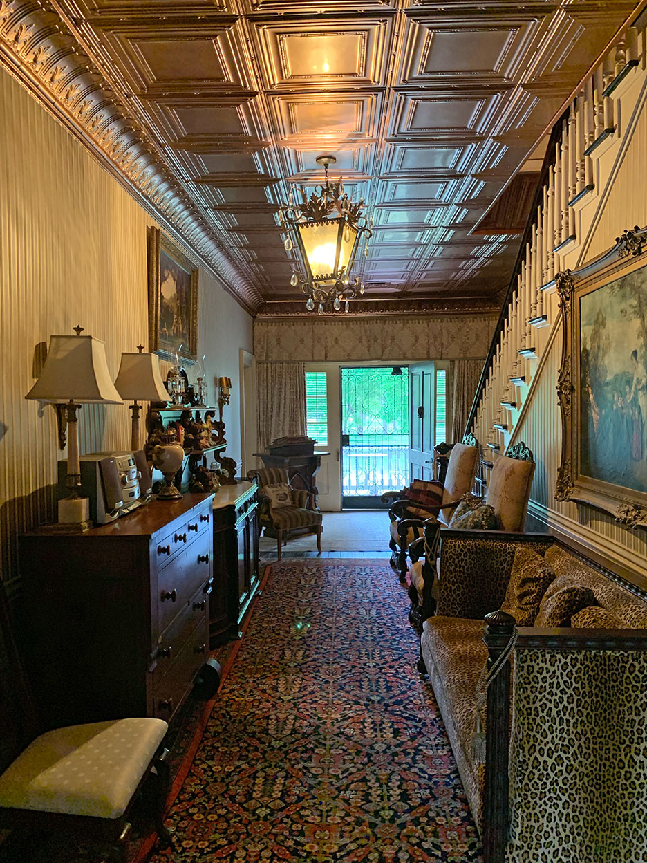 Narrow vintage hallway with patterned ceiling, rug, staircase, and antique furniture leading to a bright doorway