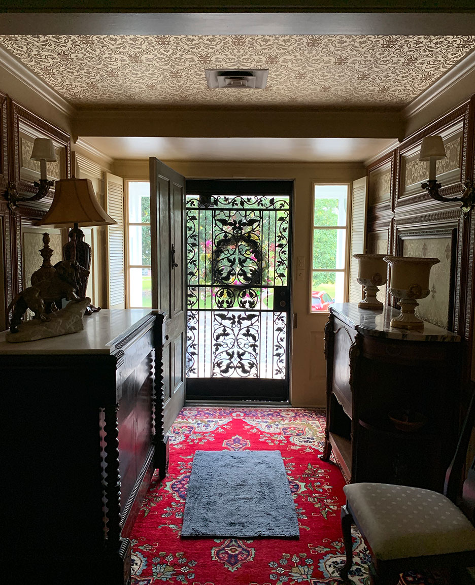 Narrow entry hall with dark wood furniture, patterned ceiling, red rug, and decorative glass front door