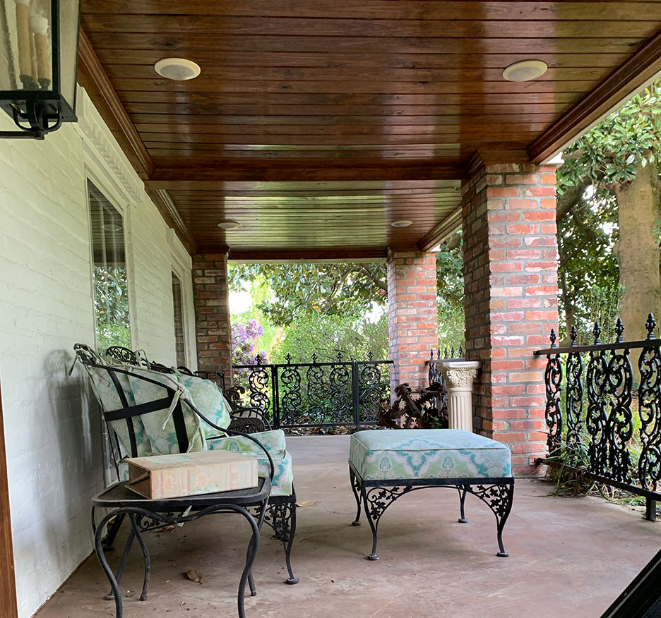 Covered front porch with wood ceiling, brick columns, wrought-iron furniture, and potted plants overlooking a yard
