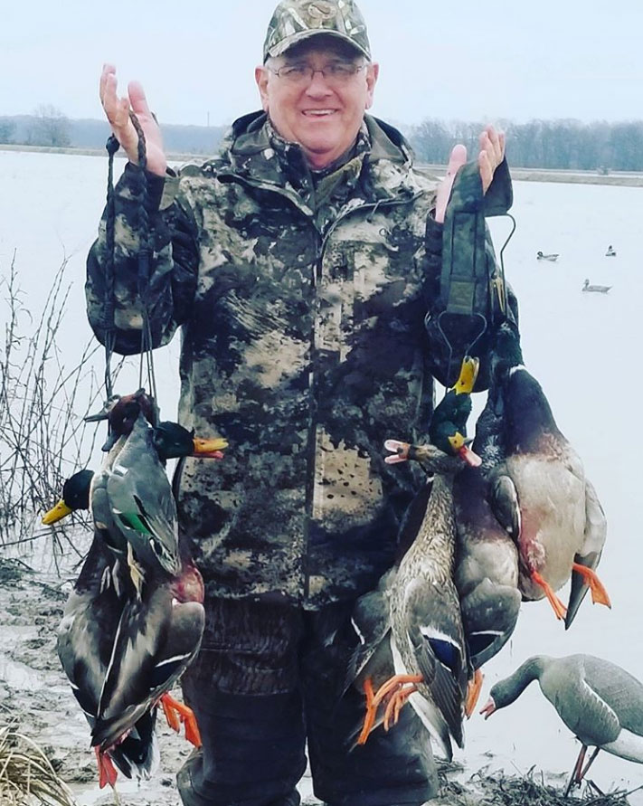 Hunter holding several ducks by a frozen lake, wearing camouflage, with birds visible nearby.
