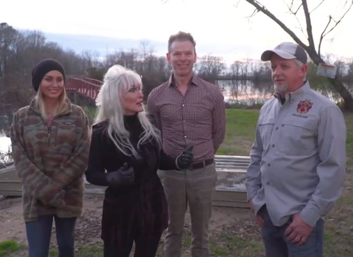 Four people standing outdoors near a lake at dusk, chatting in jackets and casual clothes.