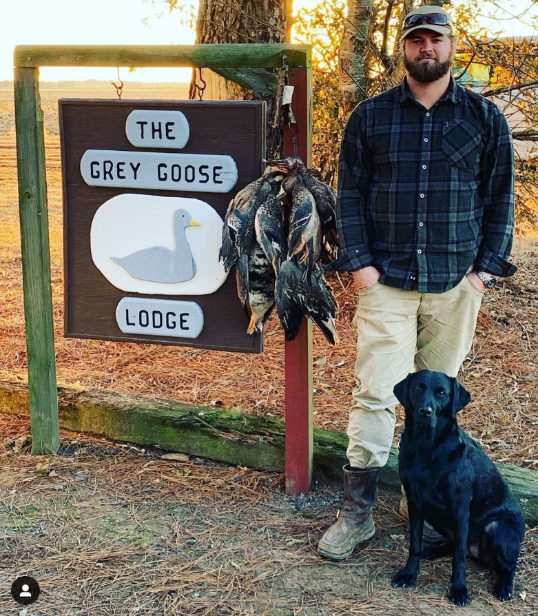 Man standing beside The Grey Goose Lodge sign, with a black dog and hanging hunting gear in a wooded area