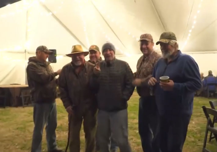 Five people standing outdoors at night in front of a lit tent, some wearing hats and holding cups