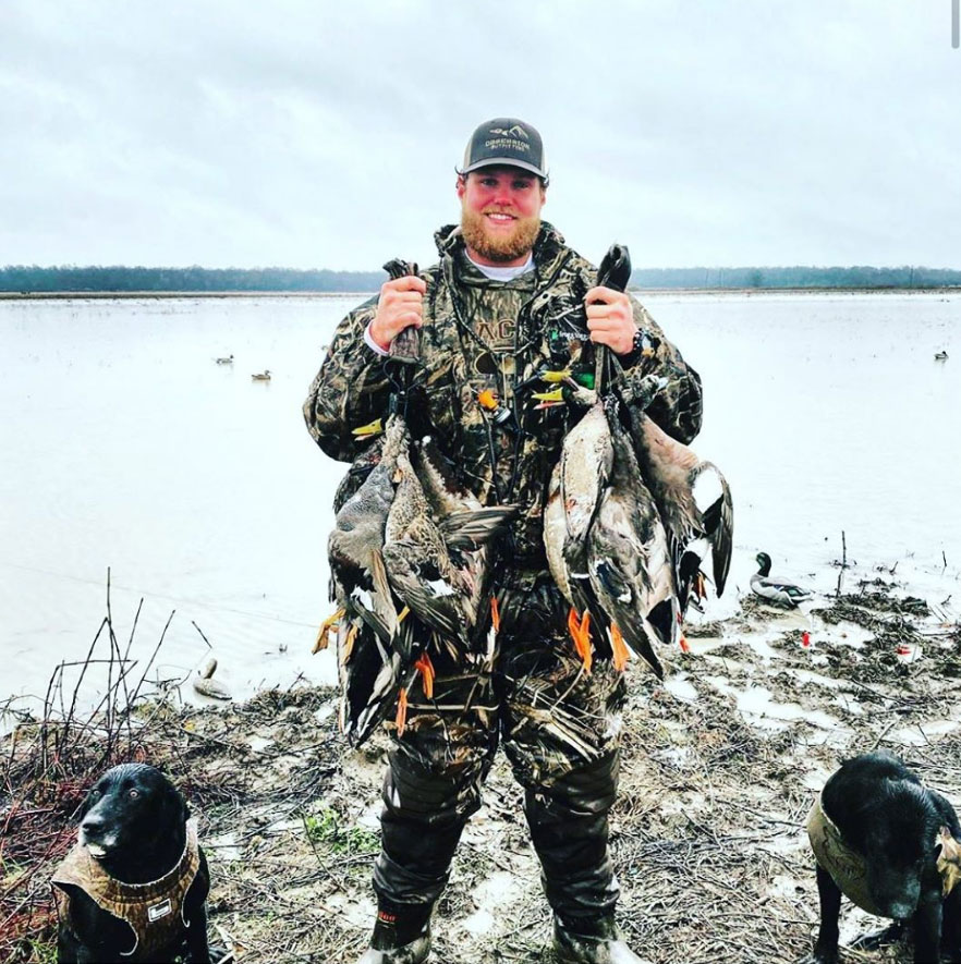 Hunter in camouflage holding several ducks by a snowy lakeshore, with two dogs beside him.