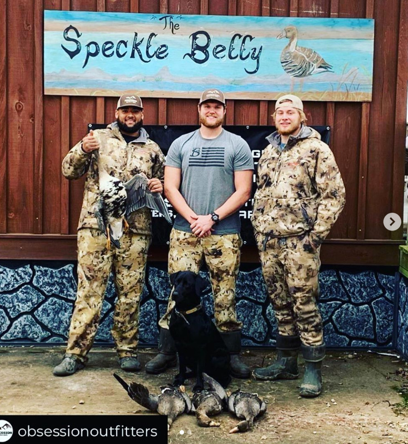Three men in camouflage holding deer beside a black dog under a Speckle Belly sign
