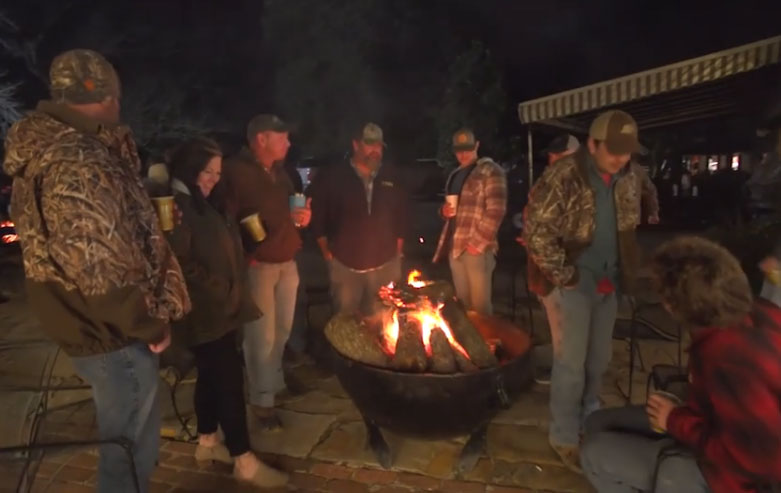 People gathered around a glowing fire pit outdoors at night, talking and holding drinks.