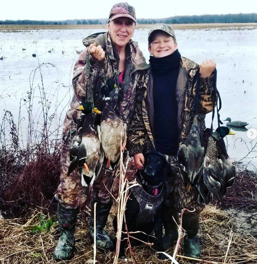 Two hunters in camouflage holding several ducks beside a snowy marsh with a black dog.