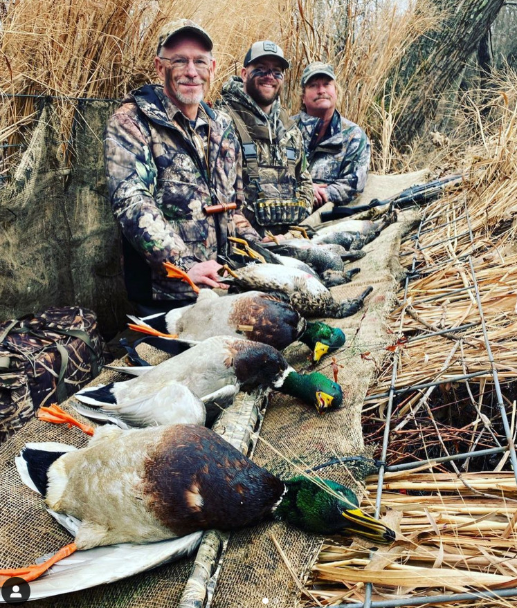 Three hunters kneel behind a row of harvested ducks in tall marsh grass.