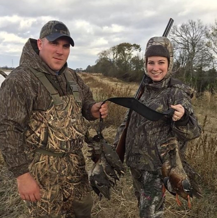 Two hunters in camouflage holding harvested birds in a grassy field under cloudy skies