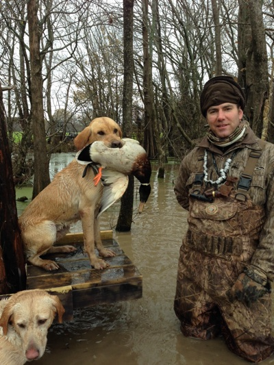 Person kneeling in flooded woods beside two dogs, one dog holding a fish, all on a raised platform